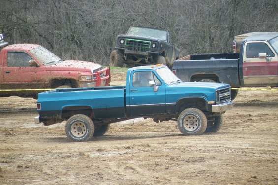 Blue and black pickup truck with off-road tires on muddy ground. Other trucks are visible in the background
