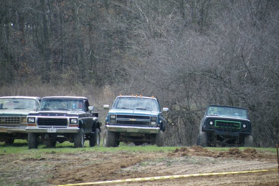 Four pickup trucks parked in a line on grassy terrain with trees in the background