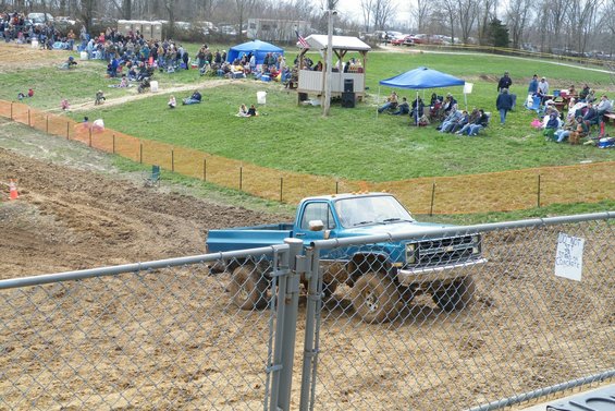 Blue pickup truck with large tires driving through mud. Sign on fence reads 'DO NOT...'