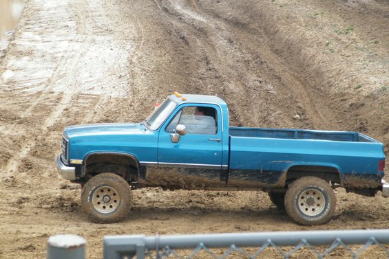 Side view of a blue pickup truck with mud splattered on its lower body, driving on a muddy track