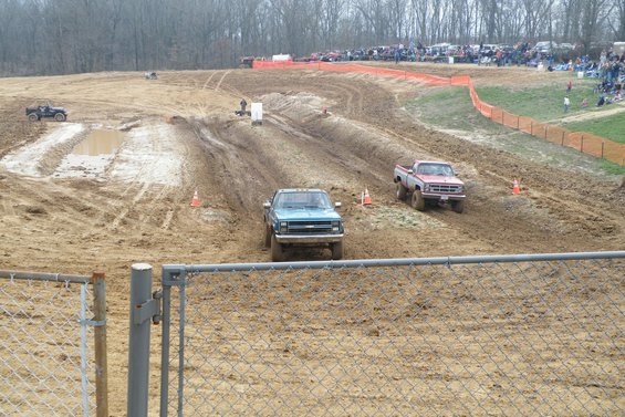 Two trucks driving through a muddy track during an event, with spectators in the background