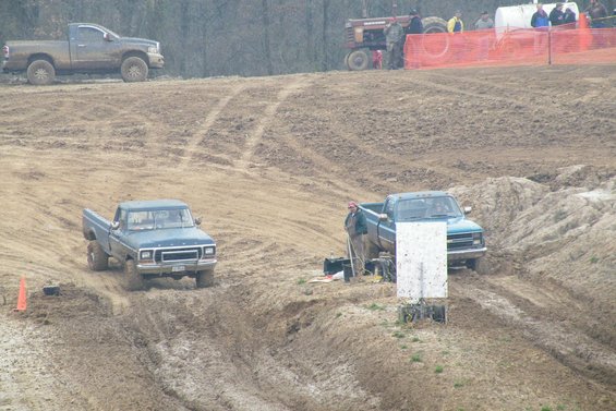 Blue Ford pickup truck driving through mud. Another truck and a tractor are visible in the background
