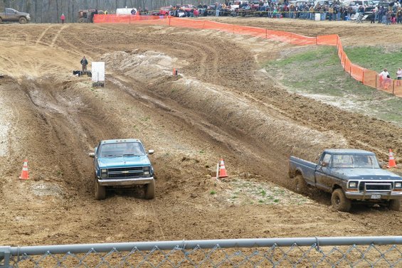 Mud bogging event with a blue Chevrolet pickup and a blue Ford pickup driving through mud