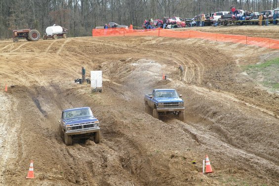 Two blue pickup trucks driving through a muddy track with spectators in the background