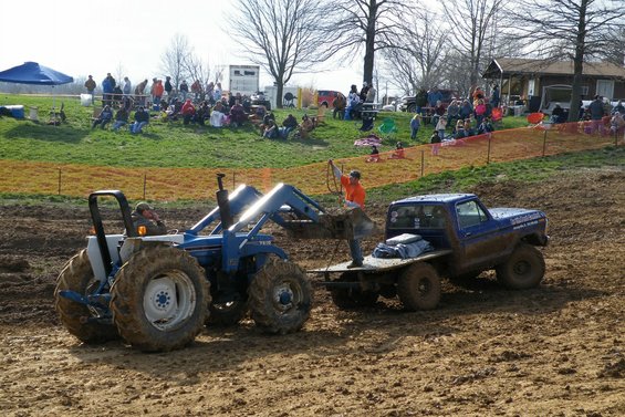 Blue tractor with front loader pulling a blue flatbed truck through mud. Spectators watch from a grassy hill