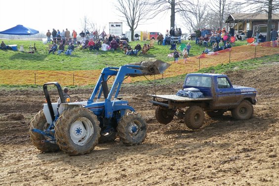 Blue tractor with loader in mud, blue flatbed truck nearby. Spectators watch from grassy hill