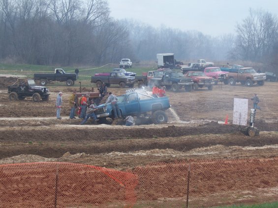 Blue pickup truck stuck in mud pit with people attempting to free it