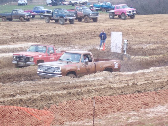 Brown pickup truck stuck in mud pit, red pickup truck behind it. Several other trucks in background
