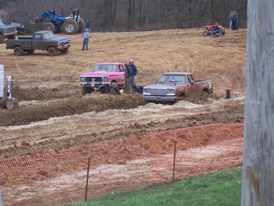Brown pickup truck stuck in mud, pink pickup truck nearby, blue tractor in background