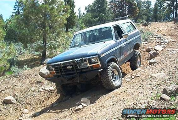 1986 Ford Bronco driving on a rocky, off-road trail with trees in the background