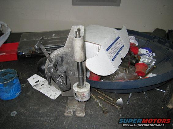 Hole saw jig with a hole saw attached, sitting on a workbench with various parts and tools in a bin