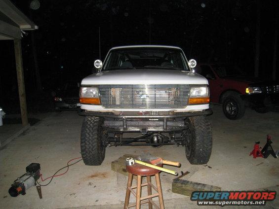Front view of a white 1996 Ford Bronco with a custom metal bumper and off-road tires. Another red truck is parked behind it
