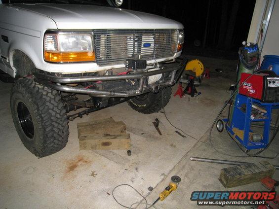 White Ford truck front end with custom metal bumper and winch. Grille has a blue Ford logo. Tools and welding equipment are in the background