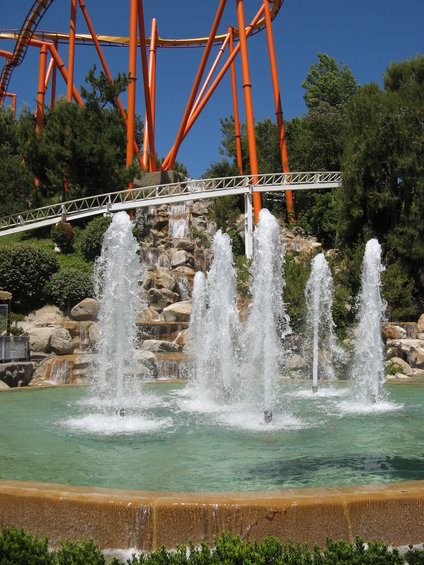 Fountains spray water in a pool with a roller coaster track in the background