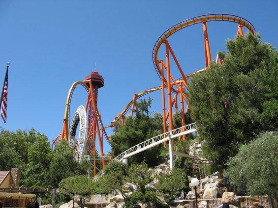 Roller coaster tracks with orange and yellow supports against a blue sky, with trees and a waterfall in the foreground
