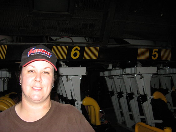 woman in a brown shirt and a black baseball cap with 'Summit Racing Equipment' logo, in front of a roller coaster track with cars numbered 5 and 6