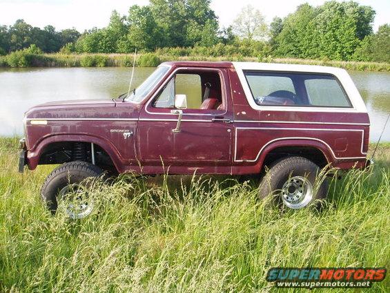 Maroon Ford Bronco with a white top, lifted suspension, and chrome wheels, parked in tall grass near a body of water