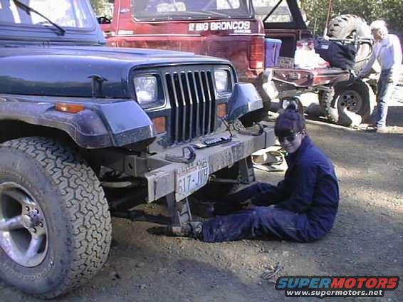 Dark green Jeep Wrangler front view, person working underneath. Red Ford Bronco in background