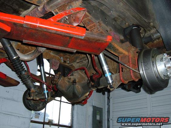 Vehicle undercarriage showing rear axle, shocks, and brake drum. Red suspension components are visible