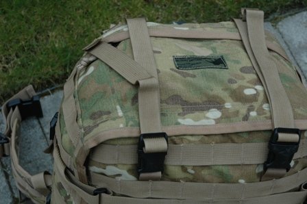 Close-up of a camouflage backpack with tan straps and black buckles. A green patch with text is visible on the front