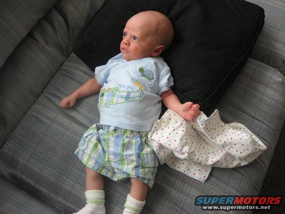 Baby lying on a couch, wearing a blue shirt with a cartoon alligator and plaid shorts