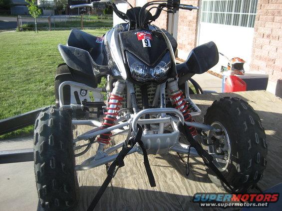 Front view of an ATV secured with straps on a wooden trailer. A red gas can and helmet are visible in the background