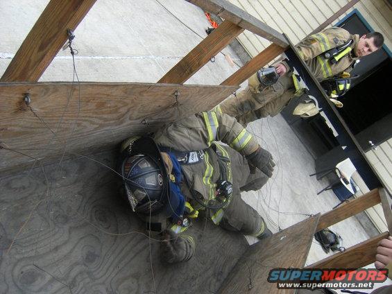 Firefighters in protective gear practice maneuvering through a wooden structure, with one firefighter on the ground and another observing from above