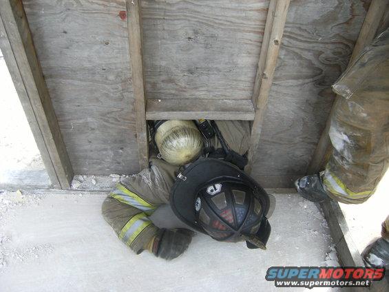 Firefighter in protective gear crawling through a wooden opening, wearing a helmet and carrying an oxygen tank