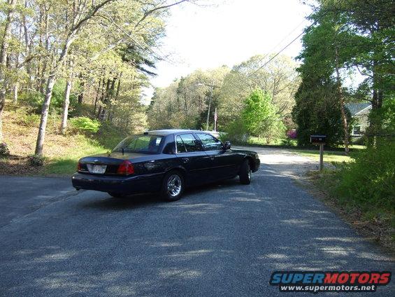 Dark sedan parked on a paved driveway with trees and a house in the background