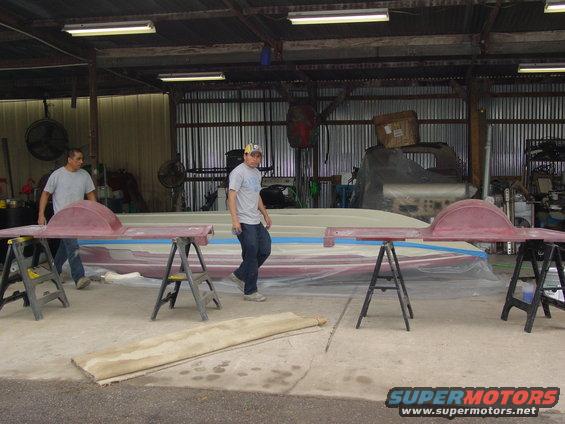 Boat hull covered in plastic sheeting, with red fender parts on sawhorses in a workshop