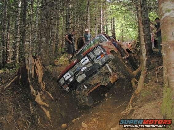 Red off-road vehicle tilted in a muddy ditch with trees around. IPF lights visible on the grille