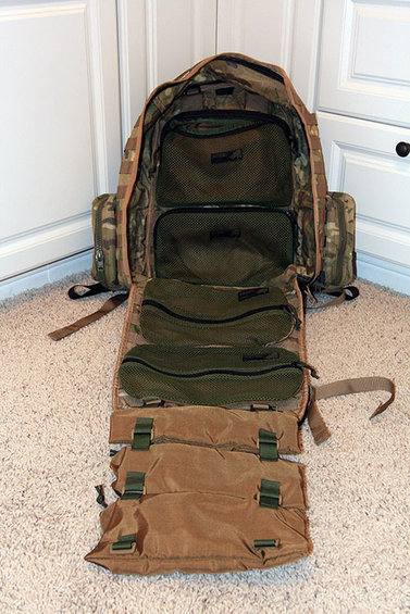 Open camouflage and tan backpack with multiple compartments and straps, lying on a carpeted floor