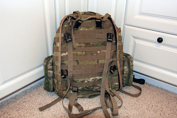 Camouflage backpack with MOLLE webbing and tan straps rests on a carpeted floor in front of white cabinets