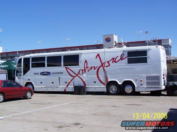 White John Force Signature Coach bus with red lettering, side view. Red car parked nearby