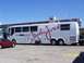 1973 Ford Mustang - A white John Force Signature Coach bus is seen parked next to a red car at a racetrack. The bus displays 'NHRA CHAMPION' and 'John Force SI…