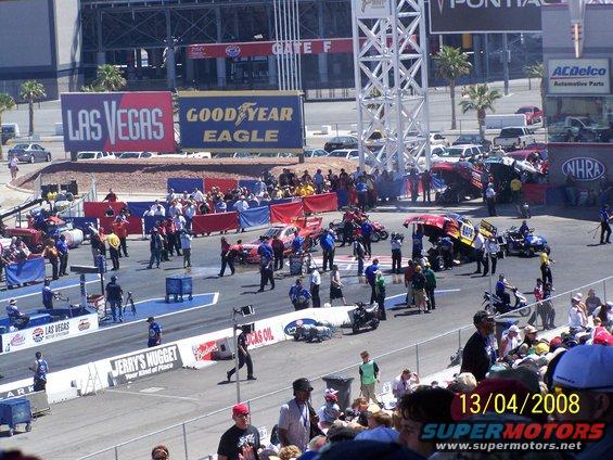 Drag strip scene with various race cars, motorcycles, and spectators under a sunny sky