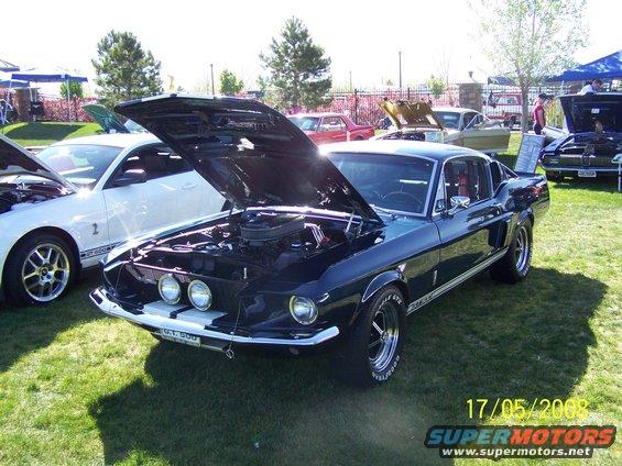 Black 1967 Ford Mustang Shelby GT500 with hood open, parked on grass at a car show