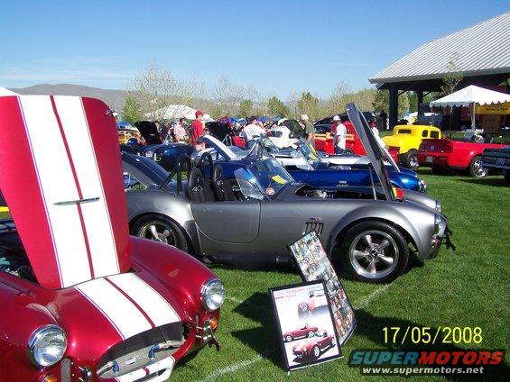Silver Shelby Cobra with hood open, parked on grass at a car show