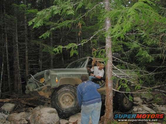 Olive green off-road vehicle with large tires, two people visible, in a wooded area