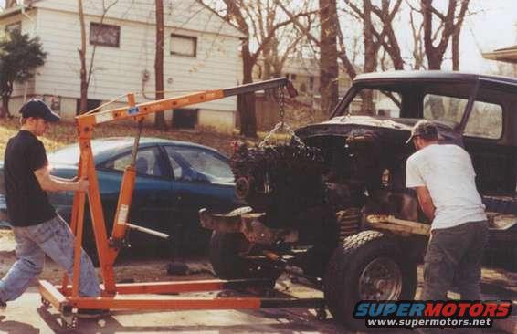 Engine hoist lifting an engine out of a dark-colored truck; another car is parked behind