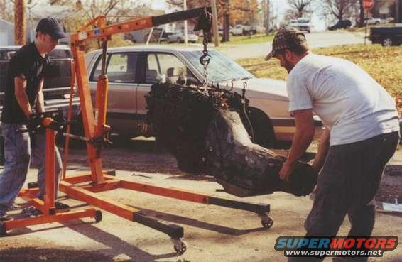 Engine hoist lifting a large engine block next to a sedan