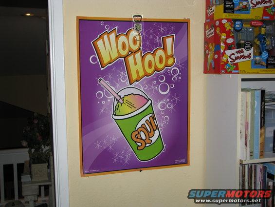 Poster with 'Woo Hoo!' text and a green cup of slushy drink. Books and Simpsons toys on shelf