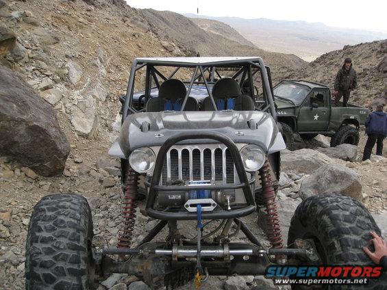 Modified Jeep with roll cage and winch, parked on rocky terrain next to a military-style pickup truck