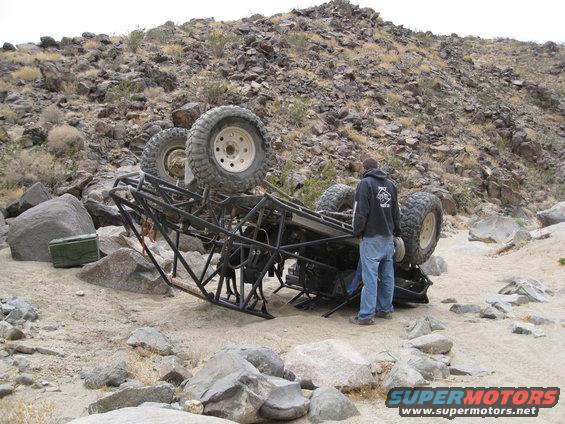off-road buggy rolled onto its side in a rocky desert landscape, a person stands nearby