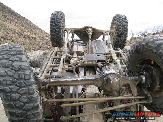 Undercarriage of a heavily modified off-road vehicle, tires visible, in a rocky, arid environment