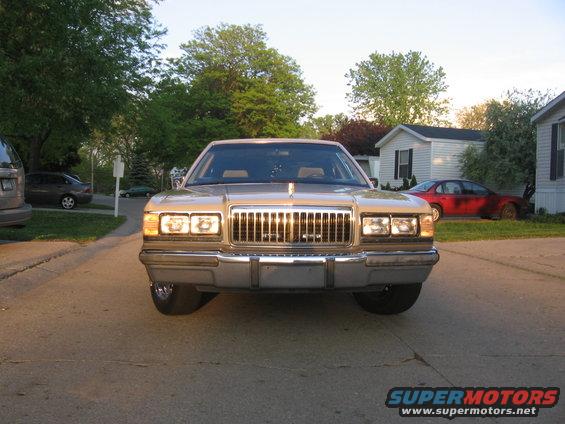 1988 Mercury Grand Marquis front view. Tan paint, chrome grille, quad headlights. Other cars and houses in background