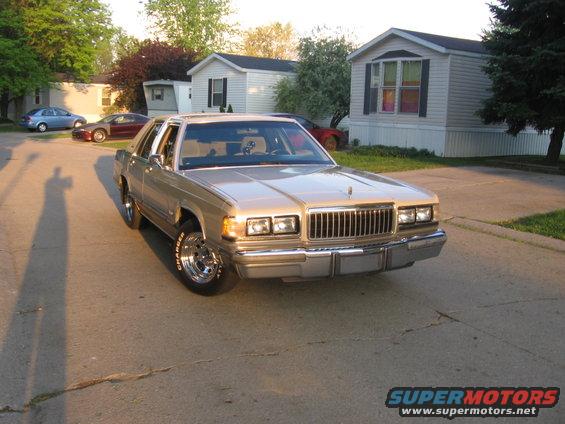 Gold 1988 Mercury Grand Marquis parked on street in front of houses. Shadow of person on left