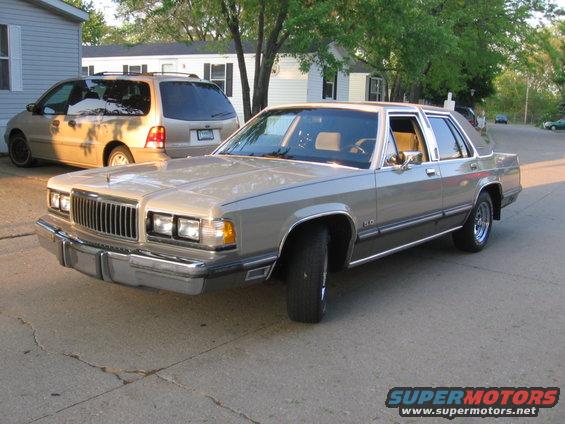 Tan 1988 Mercury Grand Marquis sedan parked on cracked asphalt, front 3/4 view, with a tan minivan behind it