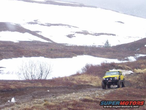 Yellow SUV with large tires on a muddy dirt road with snow-covered hills in the background