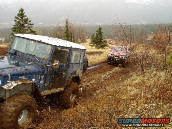 Blue off-road vehicle being towed by a red off-road vehicle on a muddy trail